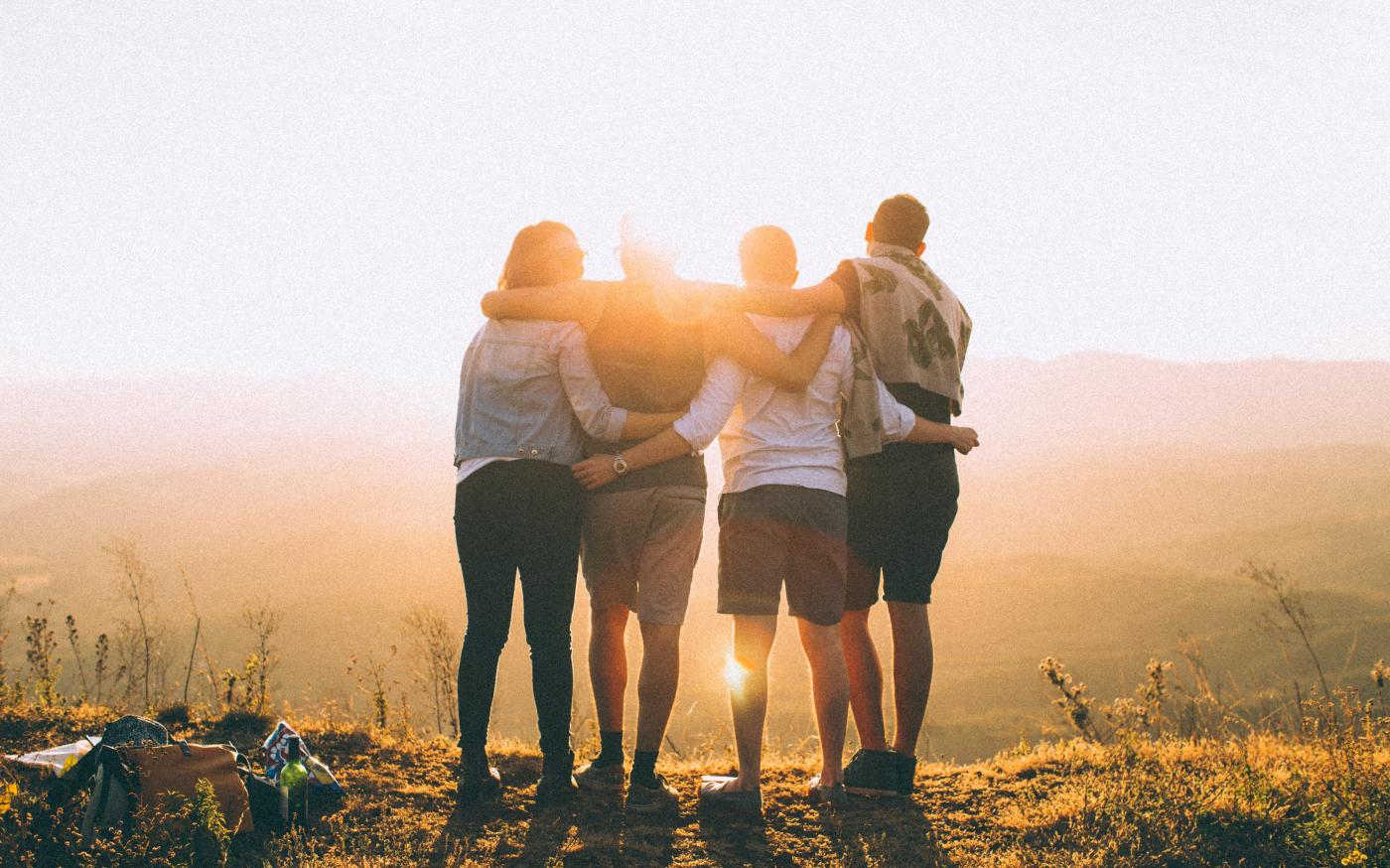four person hands wrap around shoulders while looking at sunset by Helena Lopes courtesy of Unsplash.